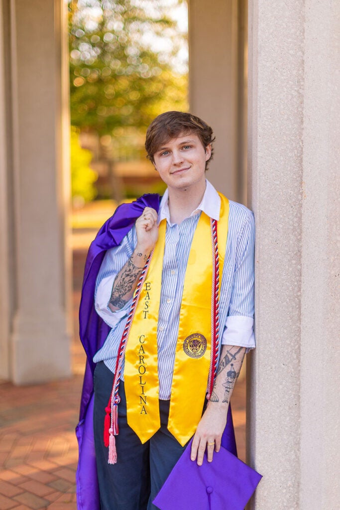 male student leaning against pillar with cap and gown in hand