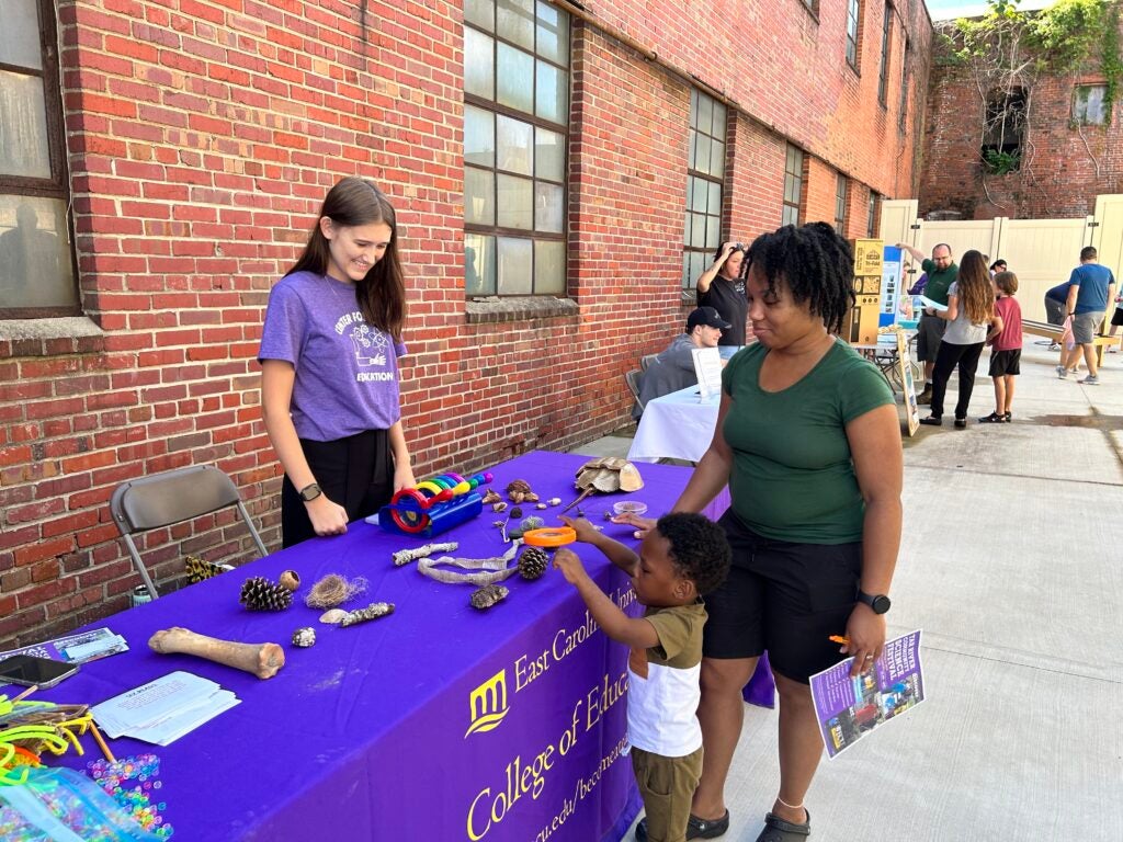 ECU student stands behind a purple table talking to an adult and child