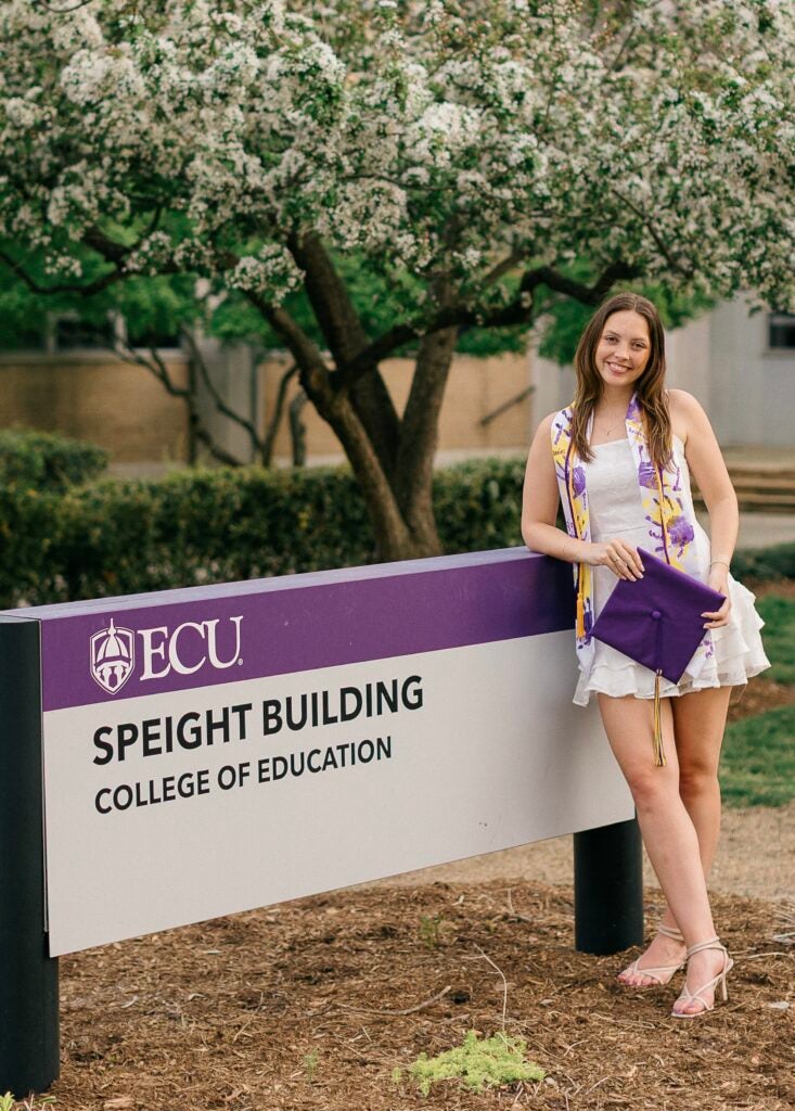 female graduate leaning against sign saying Speight Building College of Education