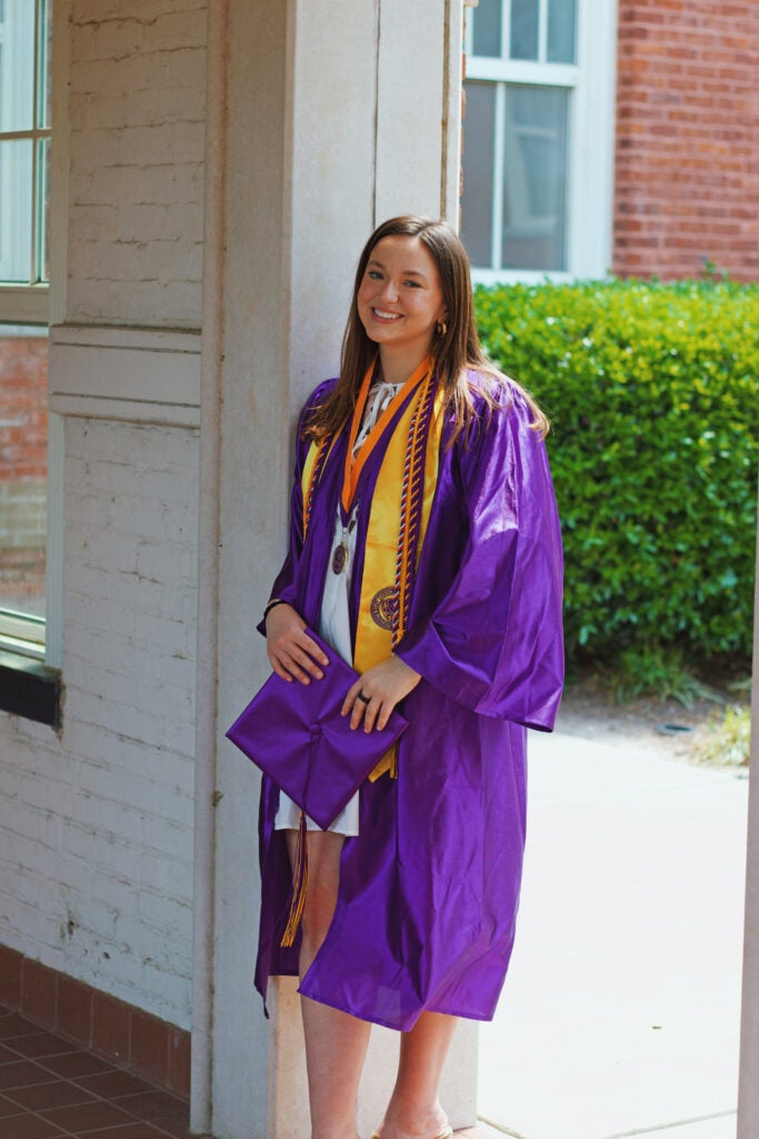 female student in purple graduation robes leaning against a white brick wall