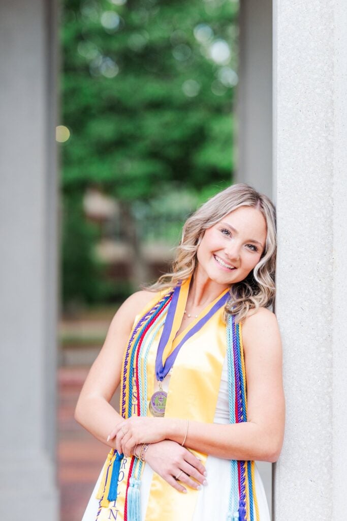 female student wearing graduation stole and cords leaning against a pillar